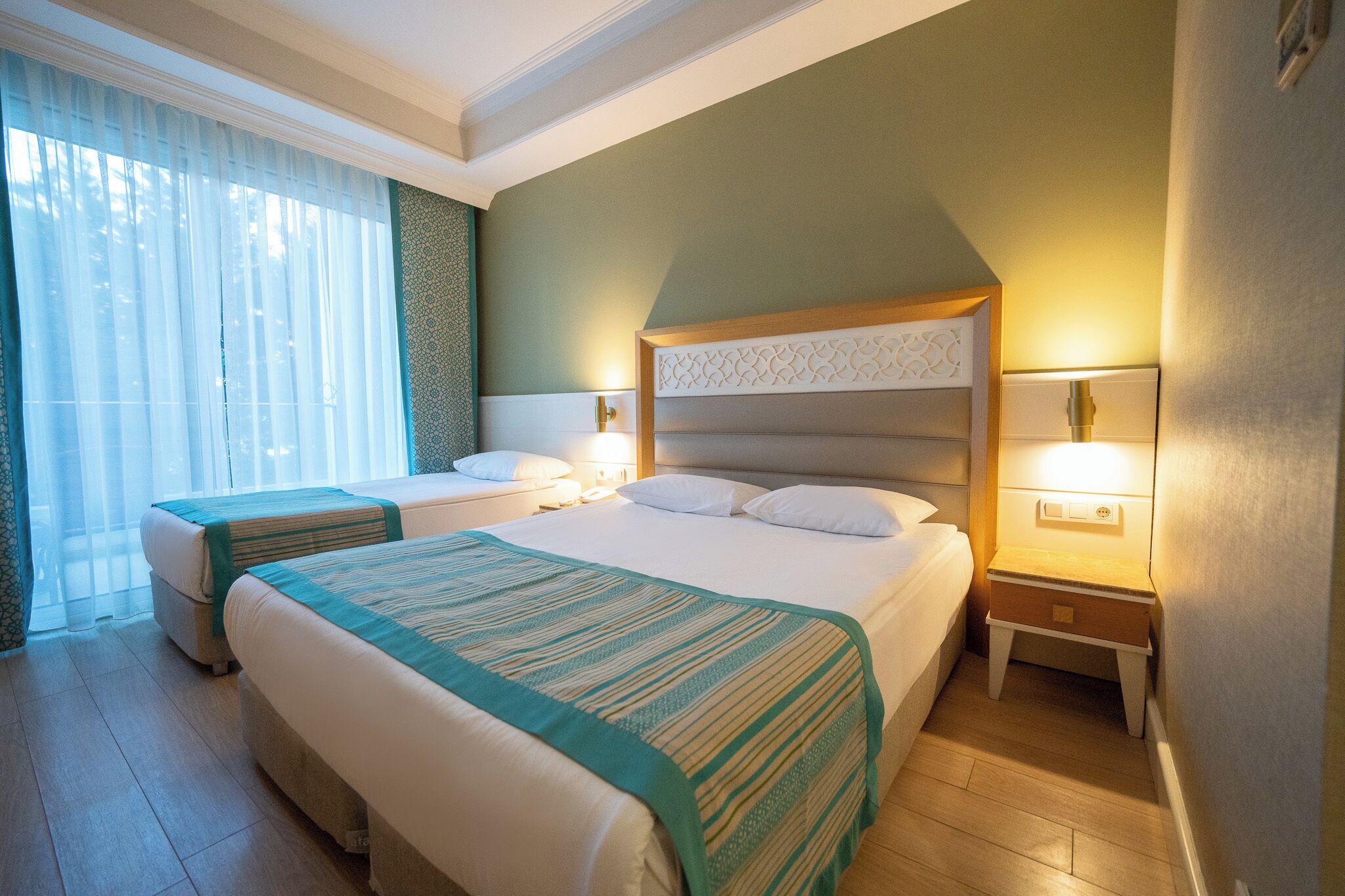 Ein Hotelzimmer mit zwei Betten, einem Nachttisch und einem Fenster mit durchsichtigen Vorhängen.