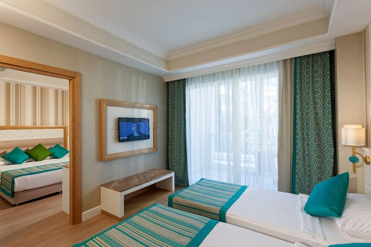A hotel room with a bed, a bedside table, a TV mounted on the wall, and a window with sheer curtains.