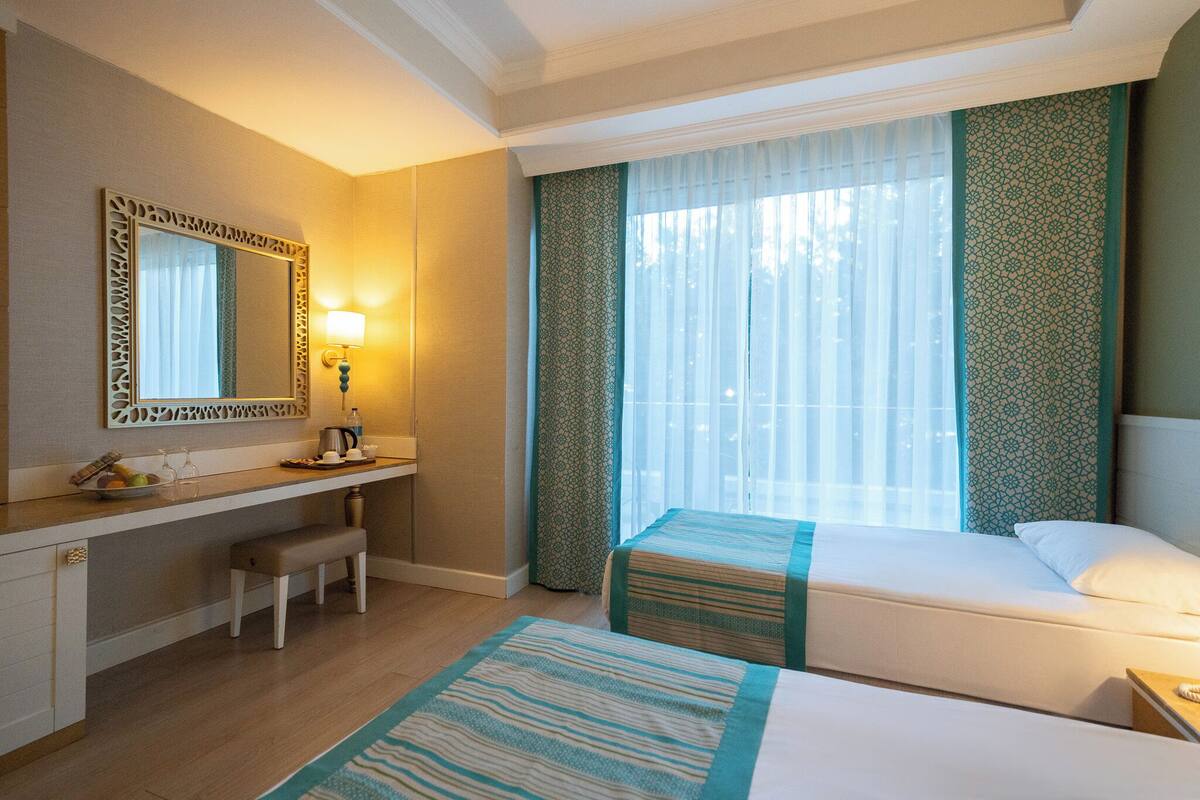 A hotel room with a bed, a bench, a desk with a lamp, a mirror, and a window with curtains.
