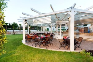 Un patio al aire libre con mesas y sillas redondas, una pérgola con un cartel y un muro de piedra al fondo.