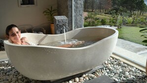A woman relaxing in a large stone bathtub with a waterfall feature, surrounded by a pebble-lined area and a view of a landscaped garden. - St Michel Wellness Hotel (Maldonado)