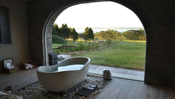 A spa room with a large bathtub, a view of a grassy landscape through an arched window, and a stone wall. - St Michel Wellness Hotel (Maldonado)