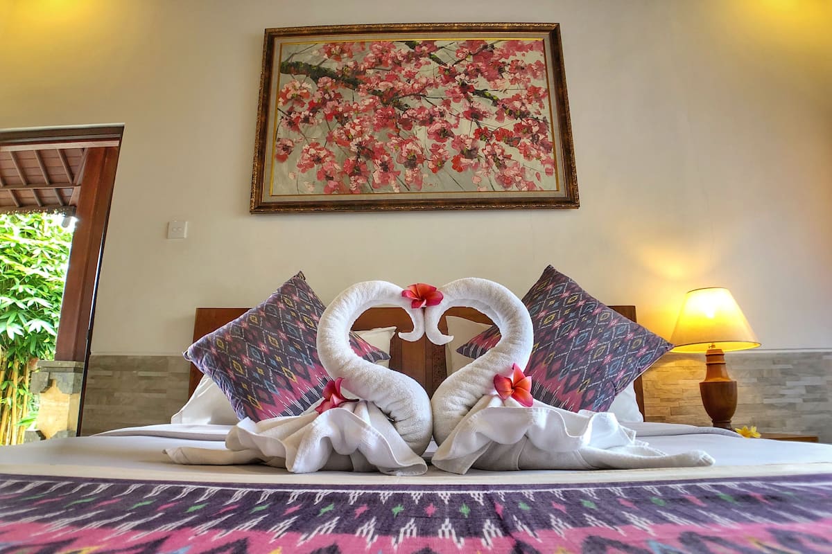 A bed with a colorful patterned bedspread, two pillows with a geometric design, and a painting of pink flowers on the wall above.