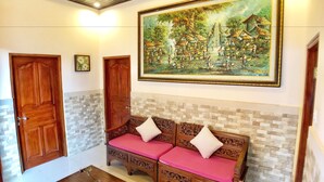 A room with a wooden bench with pink cushions, a wooden table with a glass, and a large framed painting on the wall. - Uma Dewi Guest House (Ubud)
