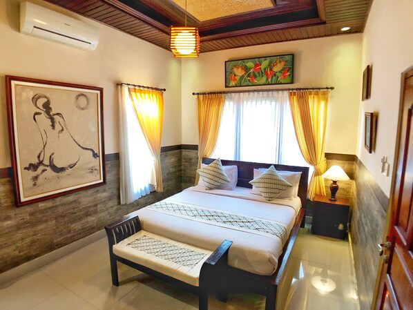 A bedroom with a bed, a nightstand, a lamp, a painting, and a ceiling fan. - Uma Dewi Guest House (Ubud)