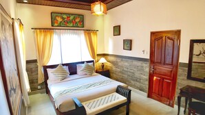 A bedroom with a bed, curtains, a wooden headboard, a bench, a nightstand, a lamp, and a door. - Uma Dewi Guest House (Ubud)