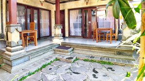 A traditional wooden porch with stone flooring, a wooden table and chairs, and a decorative stone pillar. - Uma Dewi Guest House (Ubud)