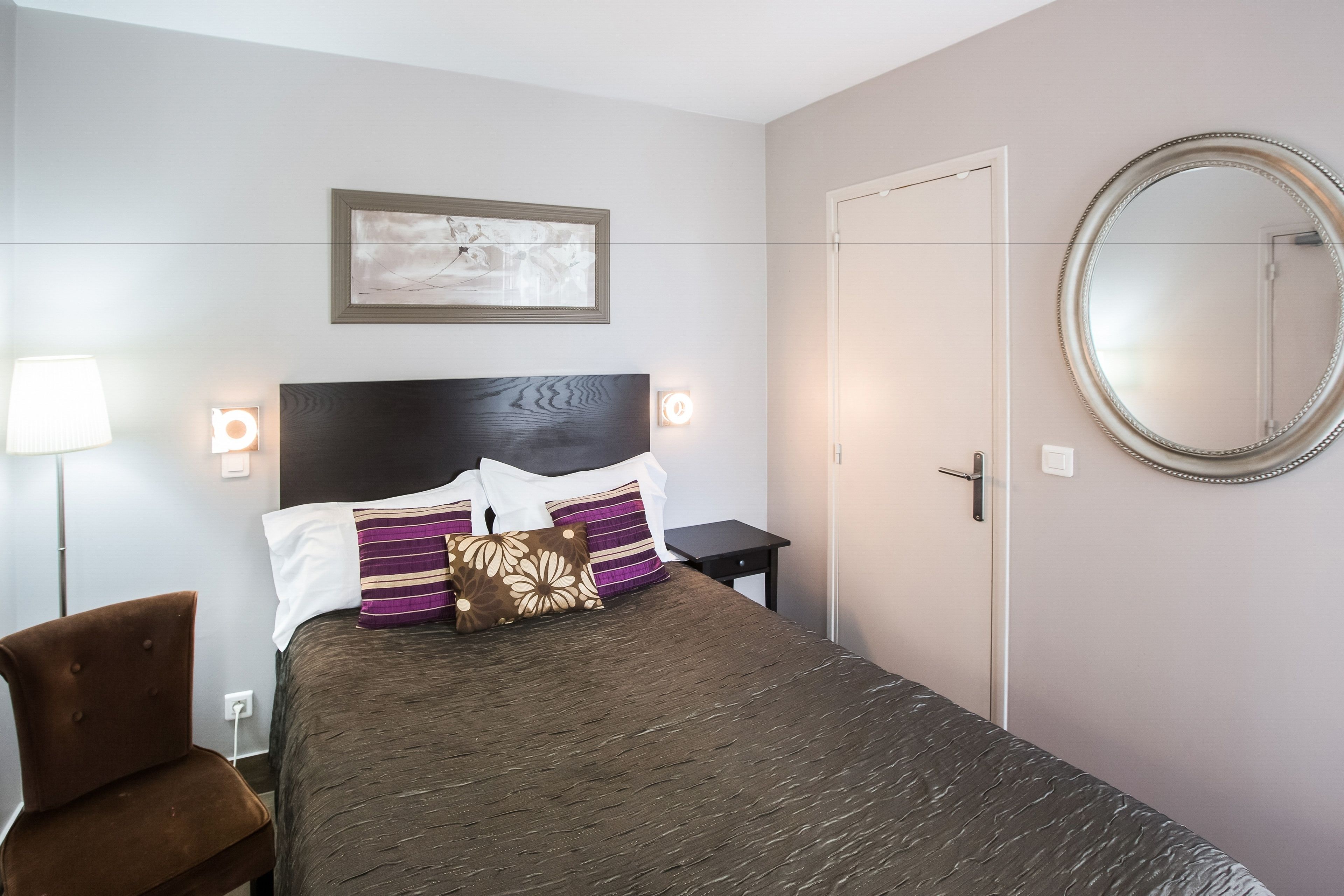 A neatly arranged bedroom with a bed, bedside lamps, a chair, a nightstand, a framed picture, and a mirror.