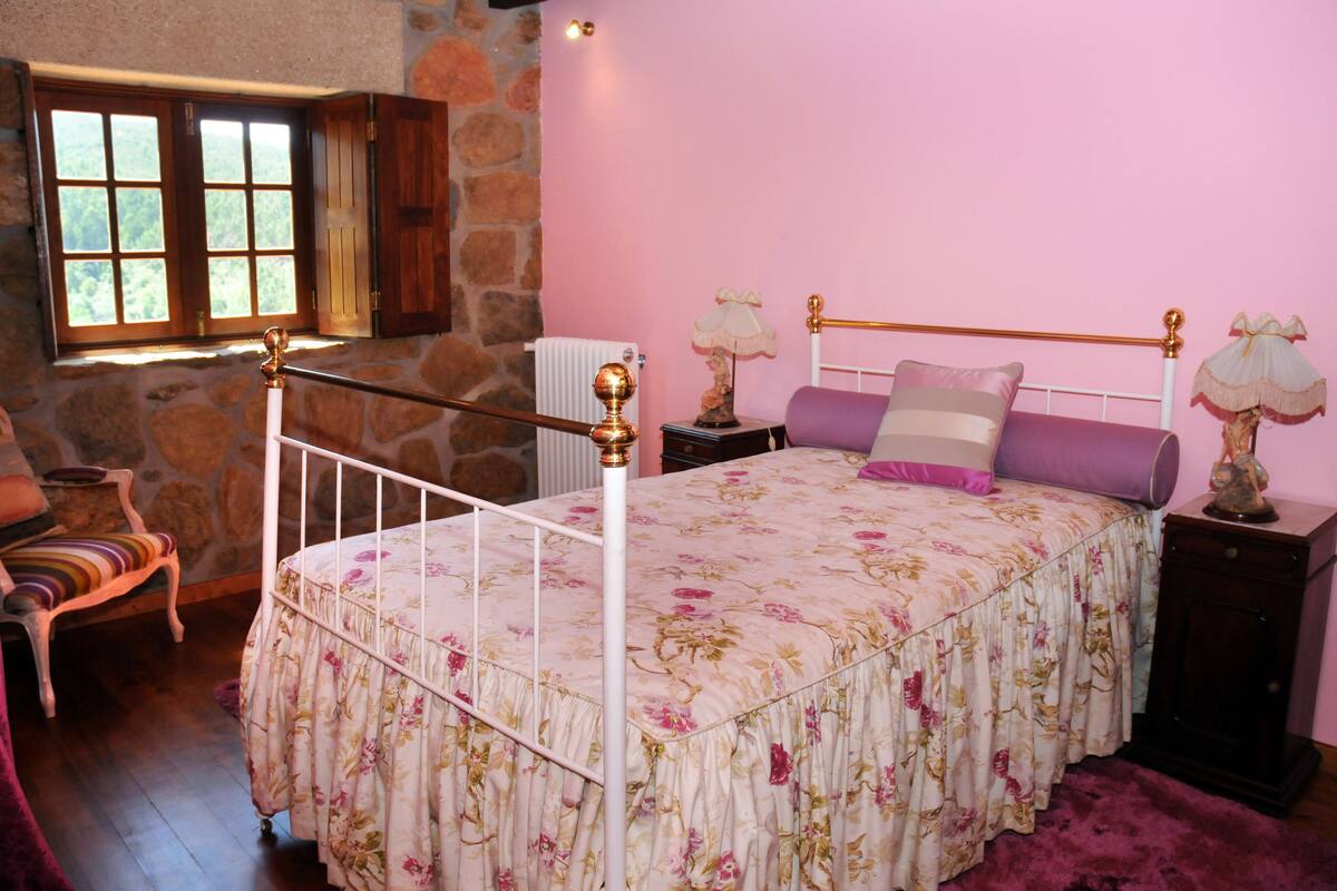 Une chambre avec un lit en métal blanc, une literie à motifs floraux, un mur en pierre, une fenêtre avec des volets en bois, une chaise avec un coussin à rayures et une table de chevet avec une lampe.