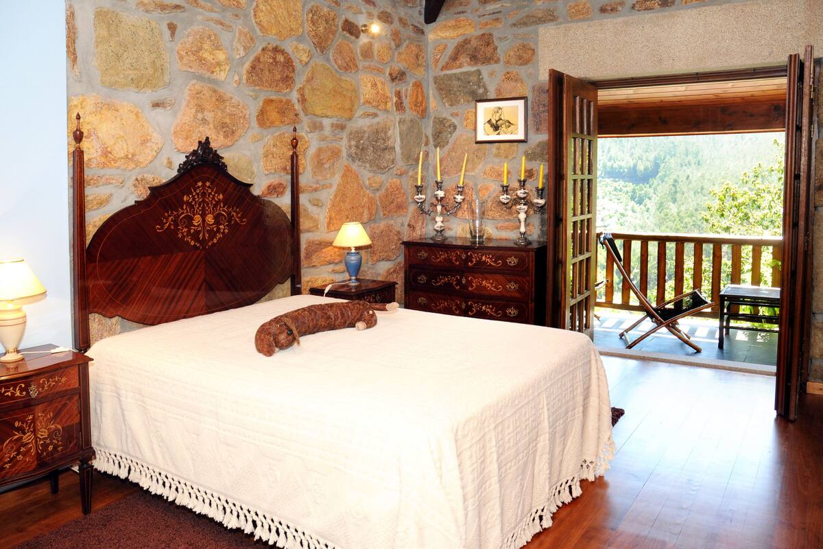 Une chambre avec un mur en pierre, un lit en bois, une table de chevet avec une lampe, une commode et un balcon donnant sur un paysage verdoyant.