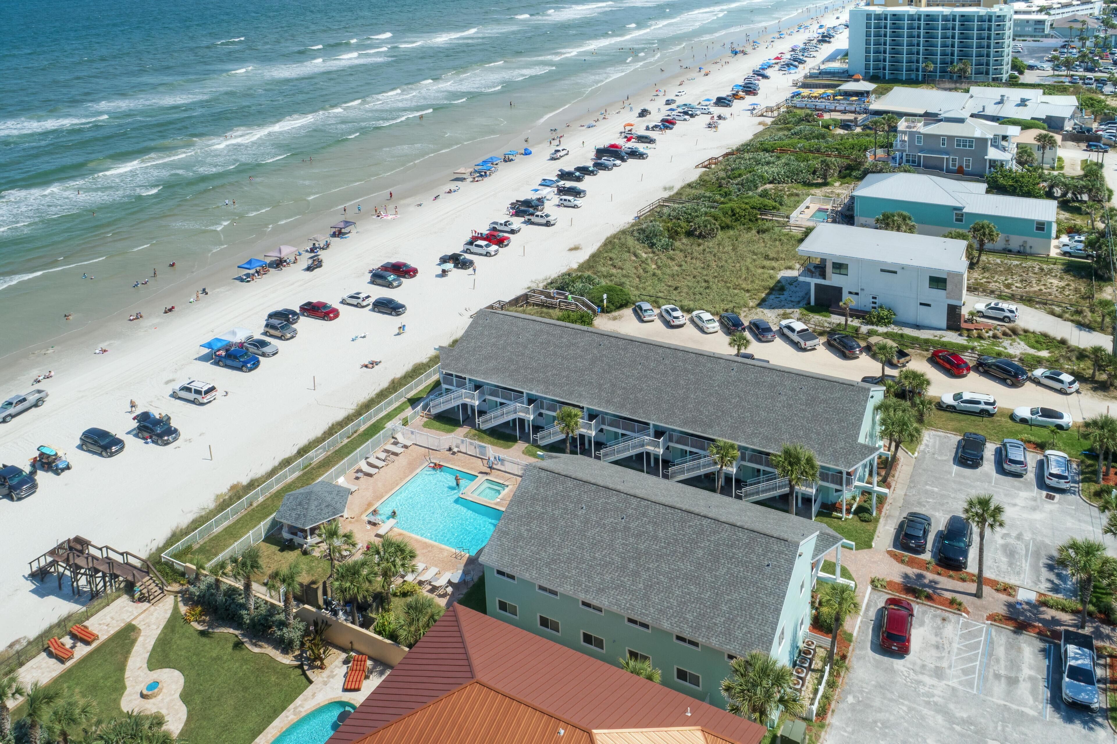 An aerial view of a coastal hotel with a beach, swimming pool, and parking area.
