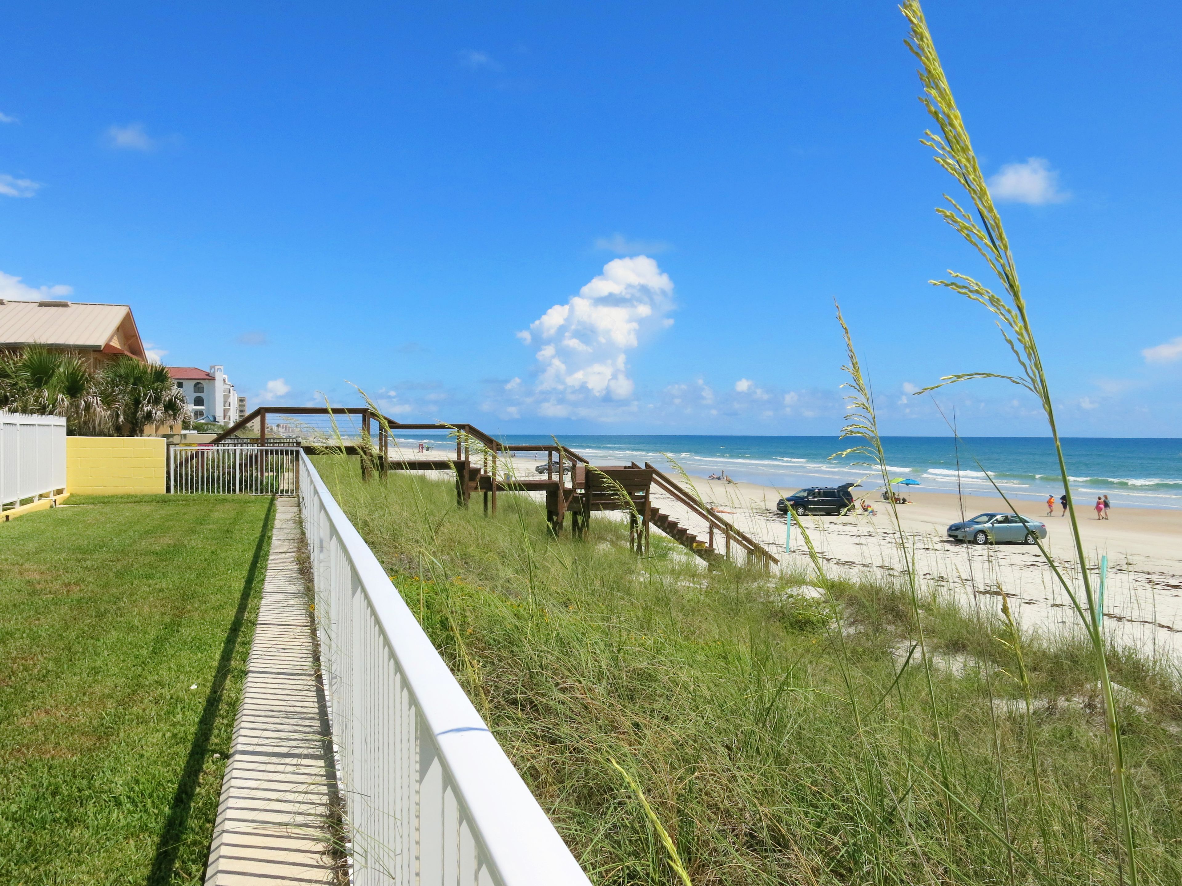 A coastal walkway leading to a beach with a clear view of the ocean and a few people in the distance.