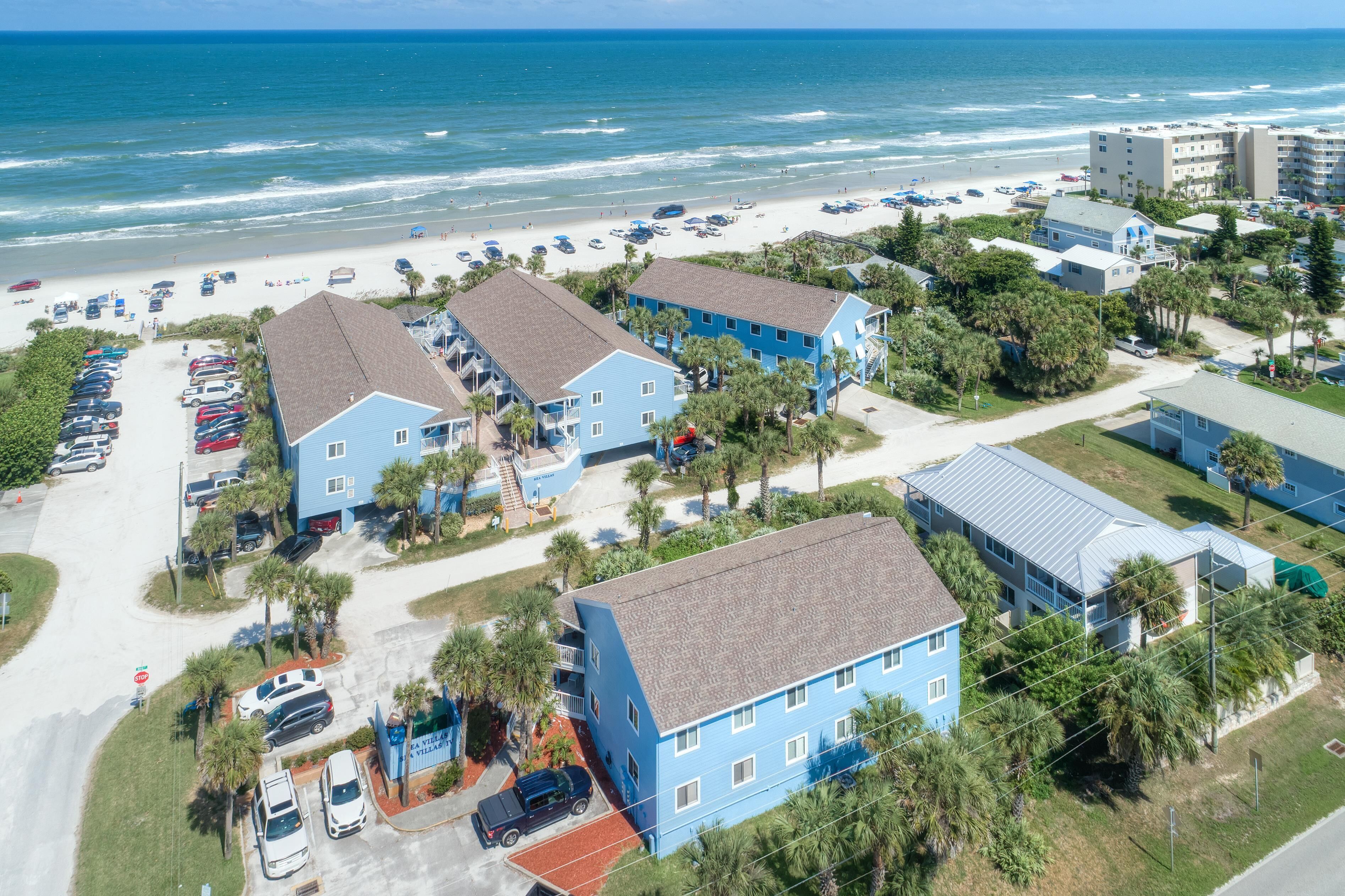 A coastal hotel with multiple blue buildings, a parking lot, and a beach view.