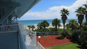 A balcony with a view of the beach, palm trees, and a red ground cover.
