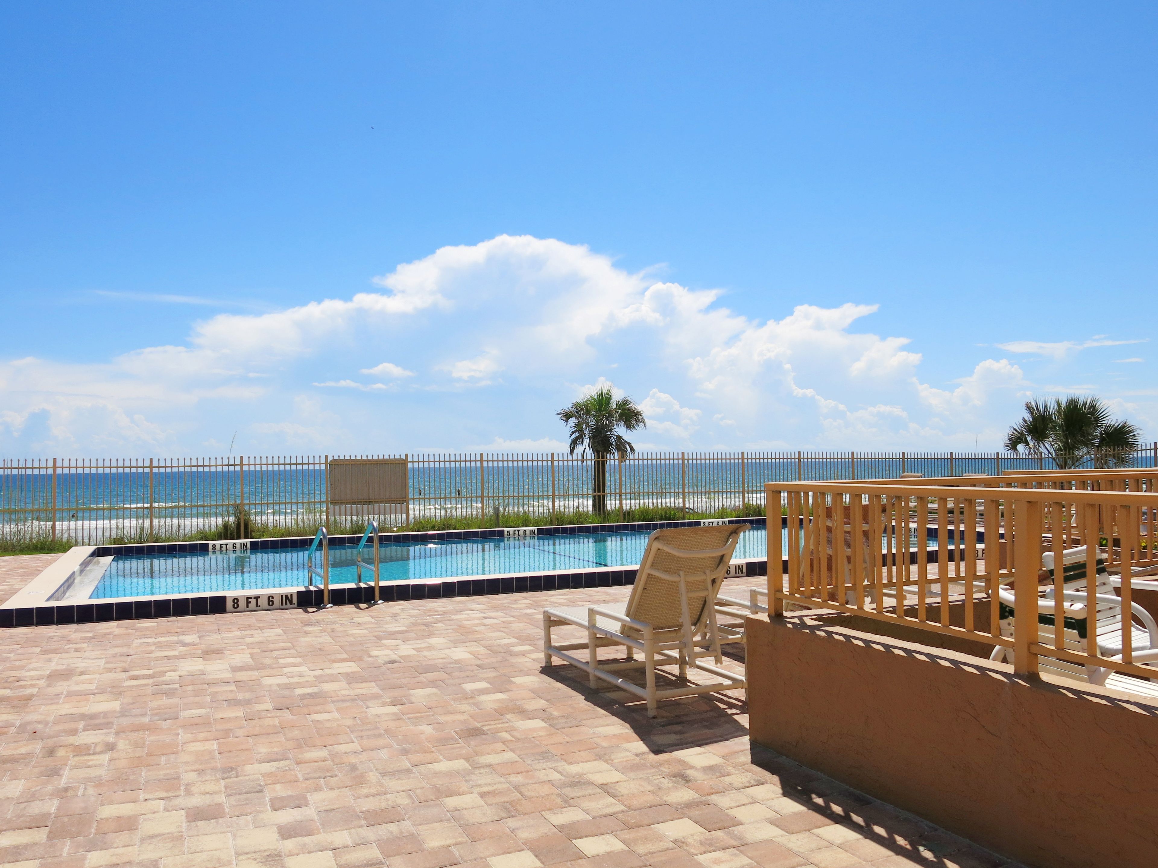 A pool area with a view of the beach, a fenced pool, and lounge chairs.