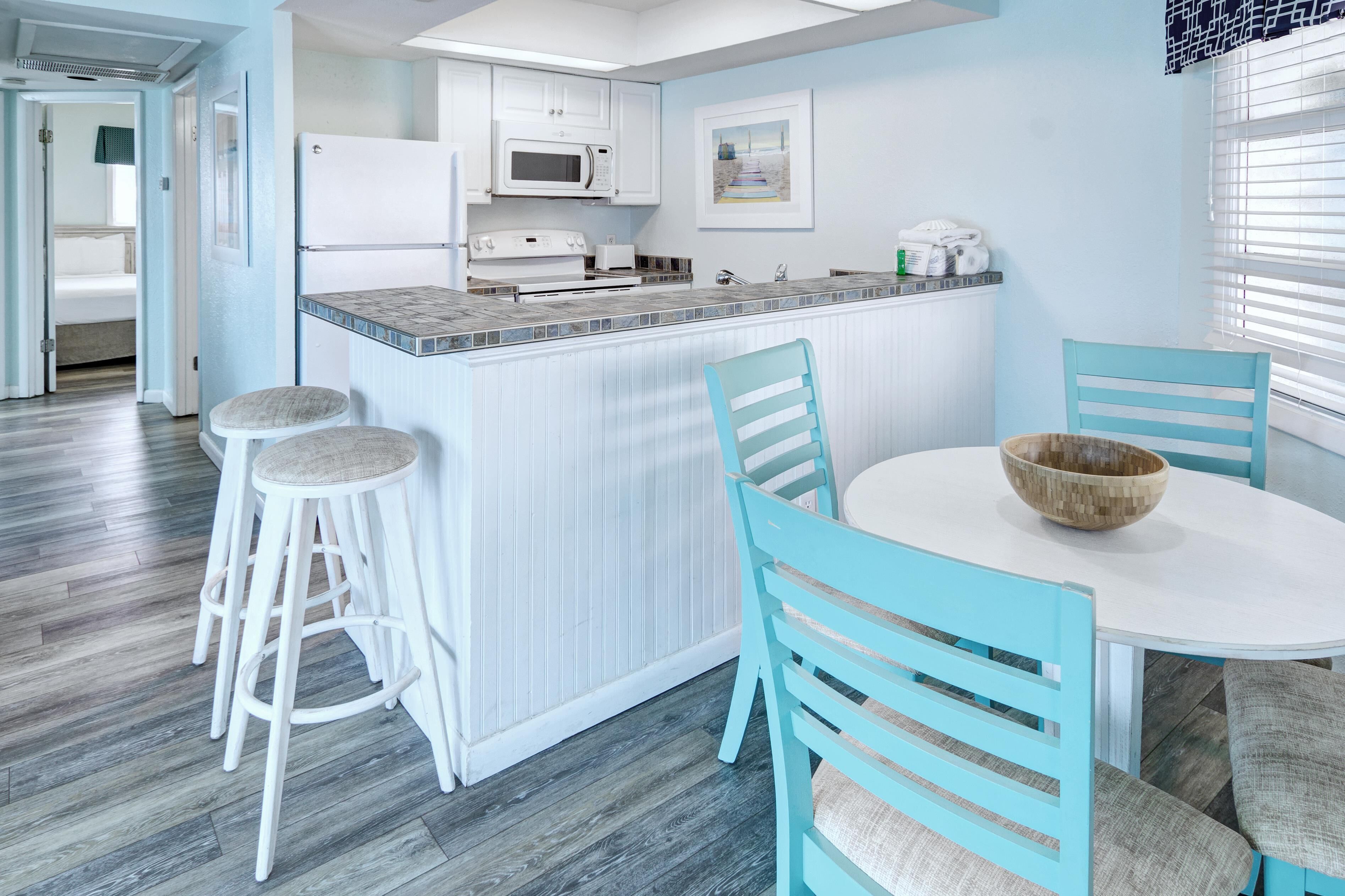 A modern kitchen with white cabinets, a microwave, and a sink. There is a dining area with a white table and blue chairs.