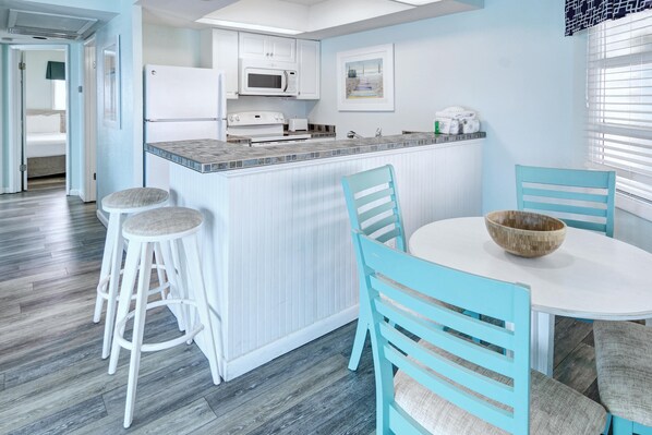 A modern kitchen with white cabinets, a microwave, and a sink. There is a dining area with a white table and blue chairs. - New Smyrna MOD Collection by Sonesta (New Smyrna Beach)
