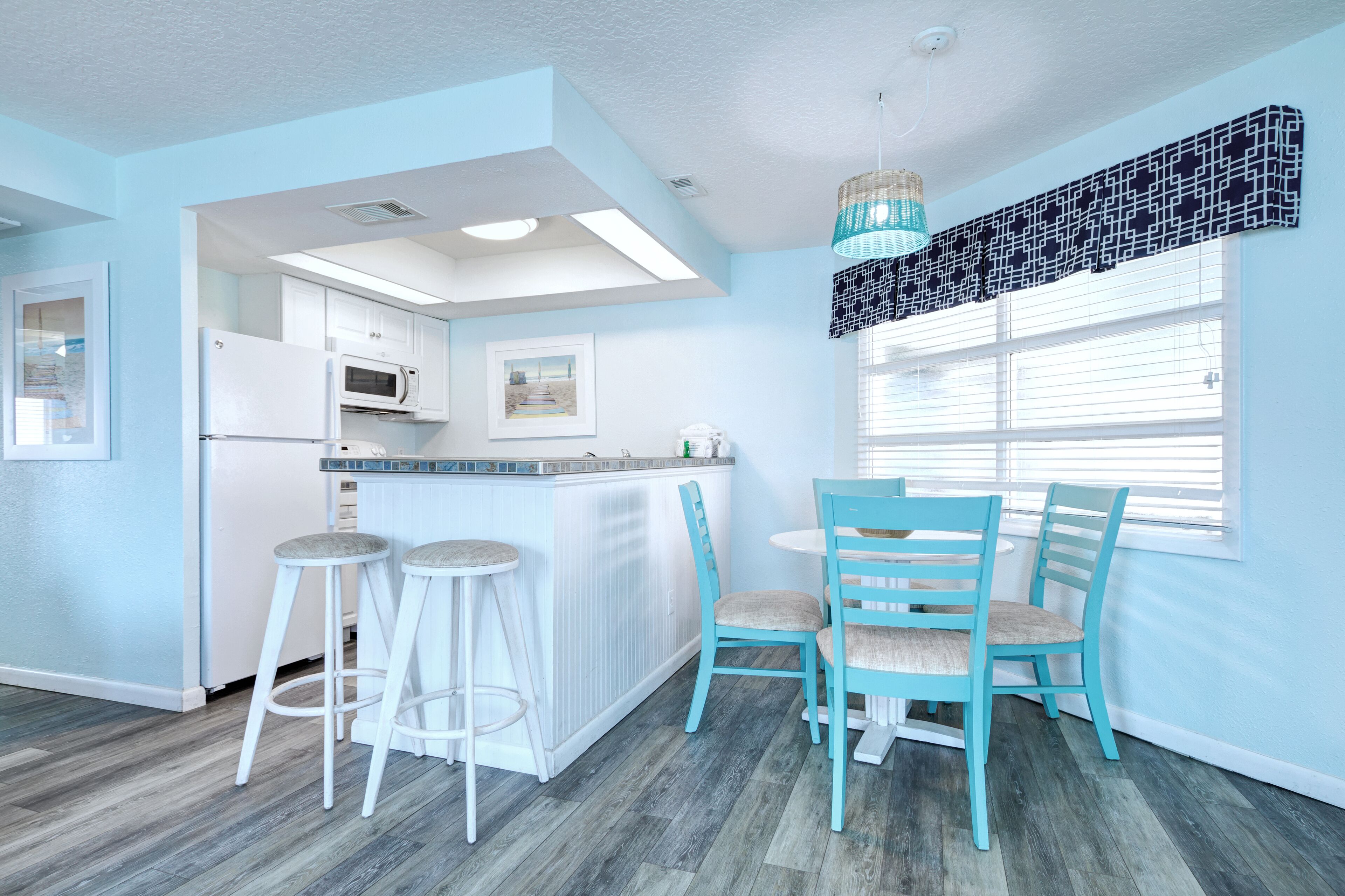 A modern kitchen with white cabinets, a white refrigerator, and a microwave. There are two white bar stools and a turquoise dining table with four chairs.