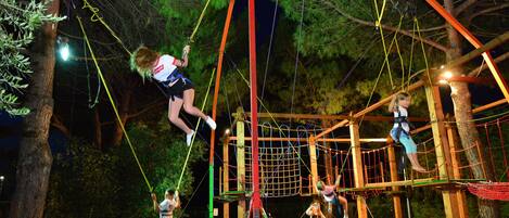 Un parque de aventuras nocturno al aire libre con puentes colgantes, trampolines y una zona de escalada con redes.