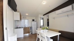 Uma cozinha compacta com armários brancos, micro-ondas e geladeira. Há uma área de jantar com mesa e cadeiras brancas, e um sofá marrom.