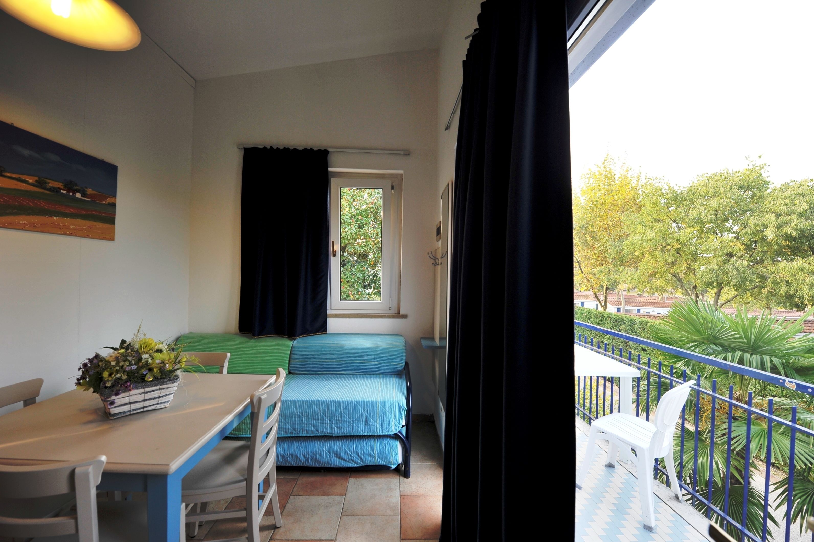 A balcony with a view of trees, a bed, a table with chairs, and a window with curtains.