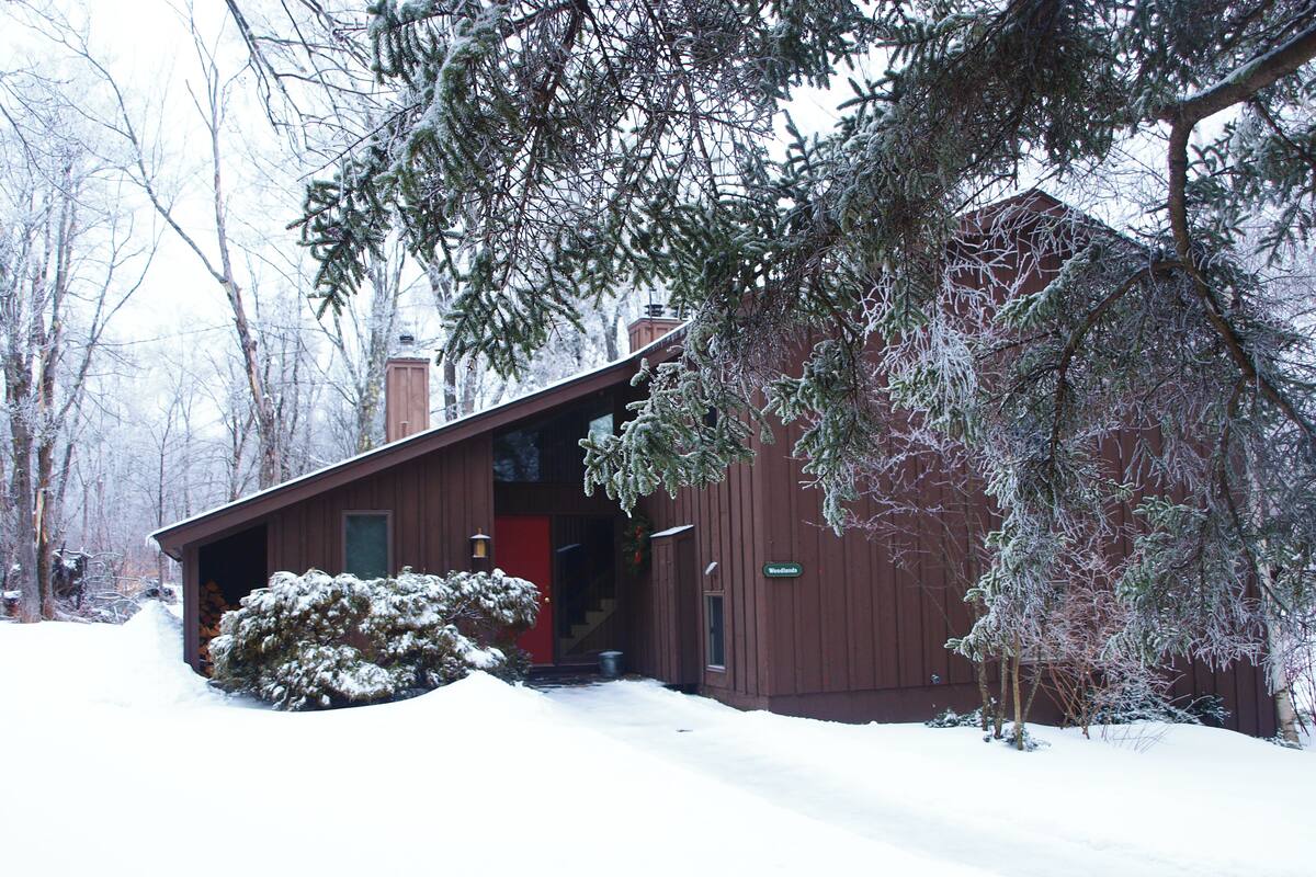 Una cabaña de madera con una puerta roja, rodeada de árboles cubiertos de nieve y suelo nevado.