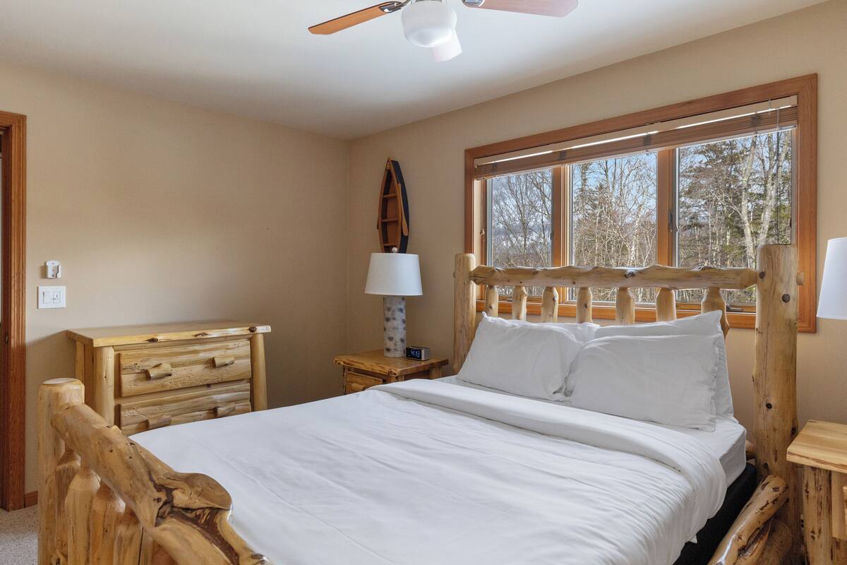 Un dormitorio con cabecera de madera, cama con ropa blanca, mesita de noche con lámpara y ventana con vista a árboles.