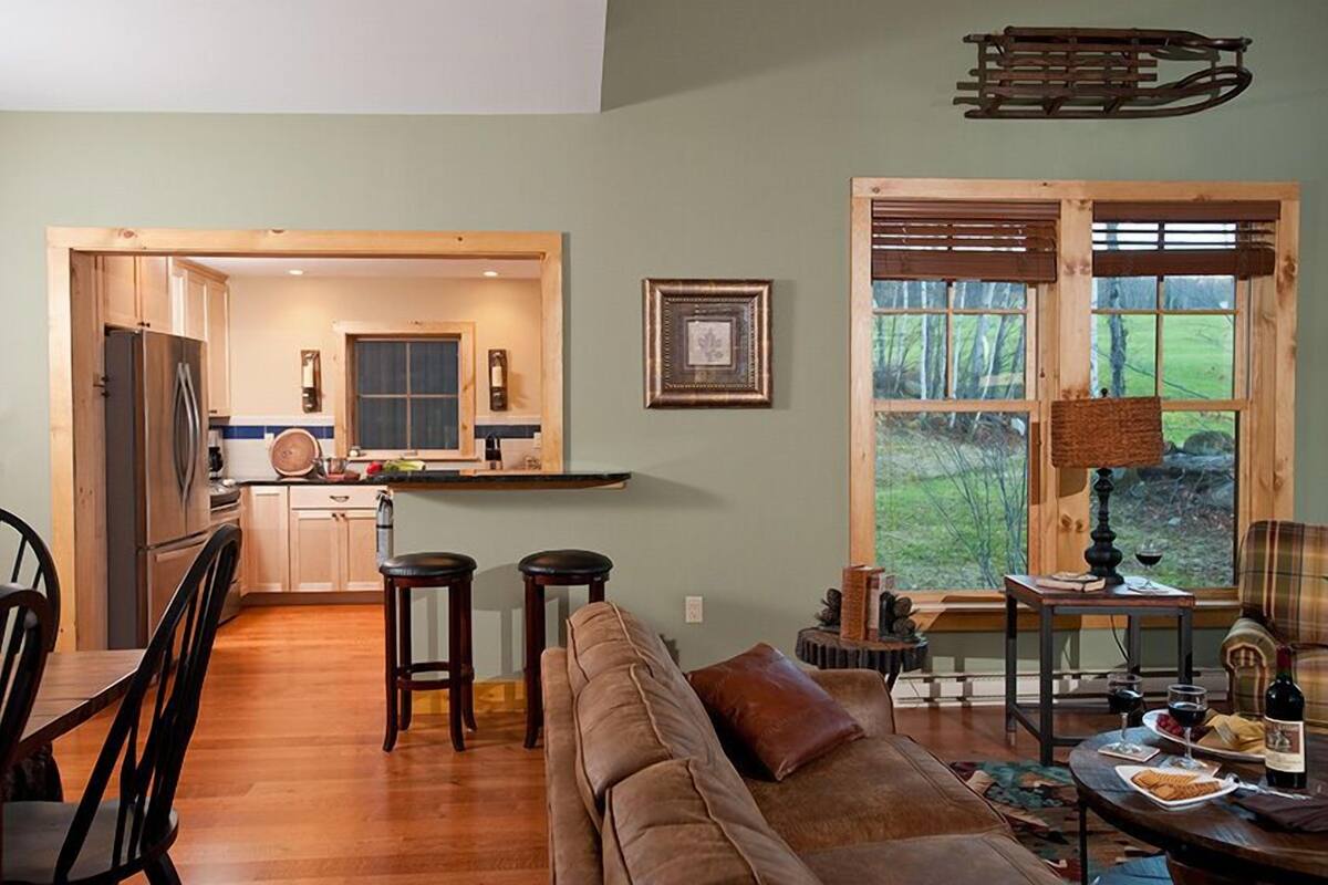 Un salón acogedor con suelo de madera, un sofá y una mesa de comedor con sillas. Se observa una zona de cocina con armarios de madera y un refrigerador. Una ventana grande ofrece vistas al exterior.