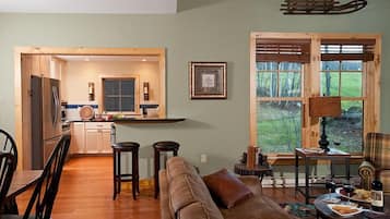 A cozy living room with a wooden floor, a sofa, and a dining table with chairs. There is a kitchen area with wooden cabinets and a refrigerator. A large window with a view of the outdoors is visible.