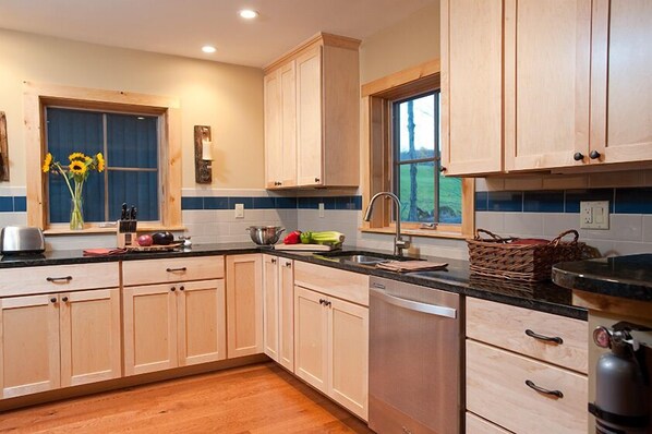 A kitchen with light wooden cabinets, a black countertop, and a window with a view of greenery.