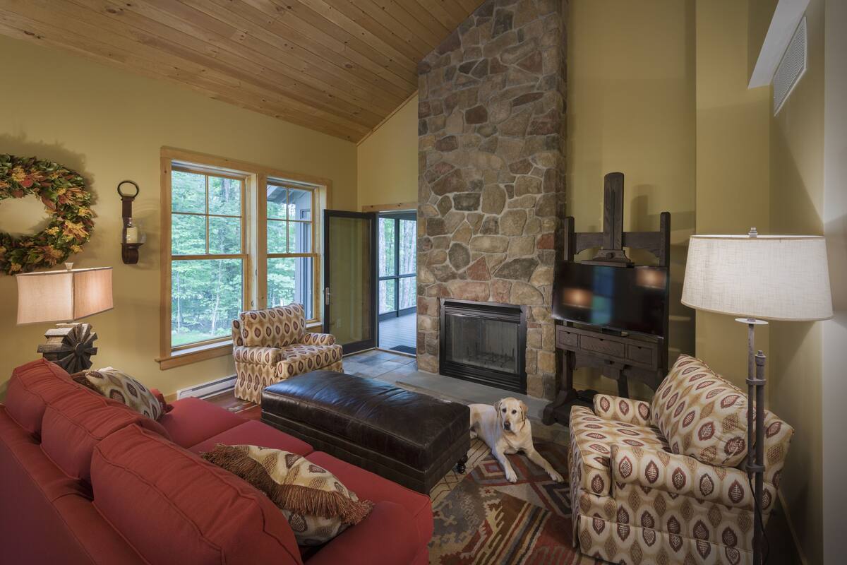 Una sala de estar acogedora con chimenea de piedra, un sofá rojo, sillones estampados y un perro.