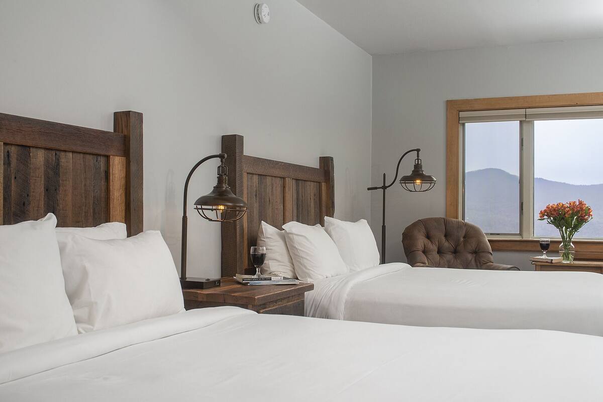Habitación de hotel con dos camas, cabeceros de madera, una mesita de noche con lámpara, una silla, un ventanal con vistas y un florero con flores.