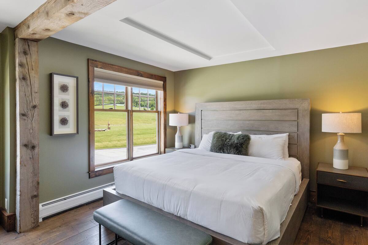 Un dormitorio con una cama grande, un banco, una mesita de noche con lámpara y una ventana con vistas a un paisaje verde.
