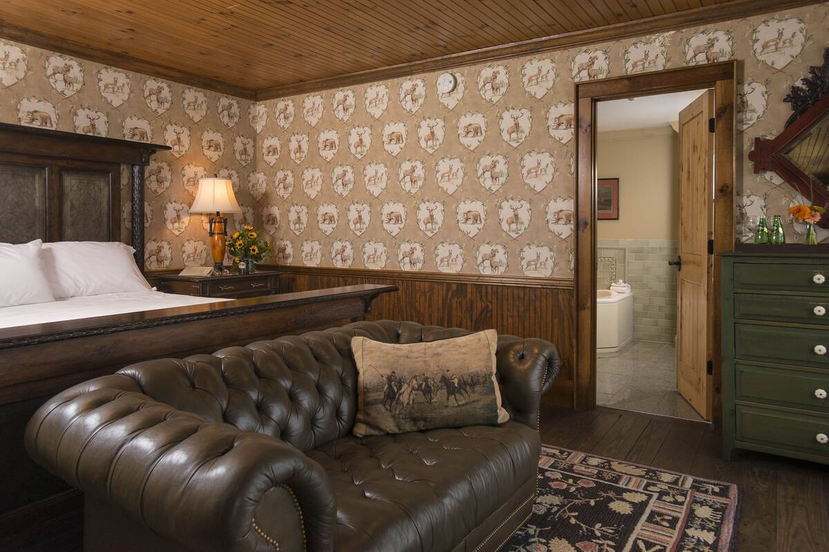 Una habitación con una cama grande, un sofá de cuero, un cabecero de madera, una mesita de noche con una lámpara y un aparador verde.