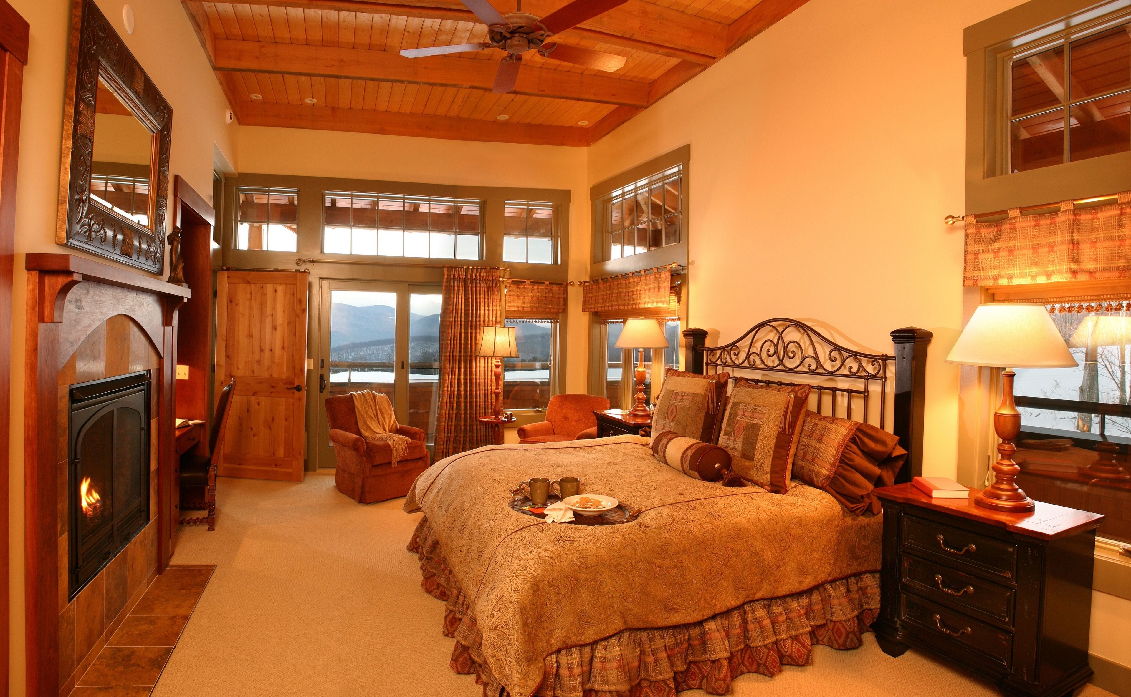 A cozy bedroom with a fireplace, a large bed with multiple pillows, a nightstand with a lamp, and a view of mountains through the windows.