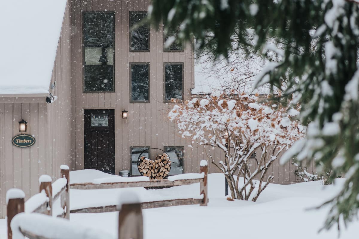 Un edificio con techo nevado y ventanas, cerca hay una valla de madera y un árbol cubierto de nieve en sus ramas.