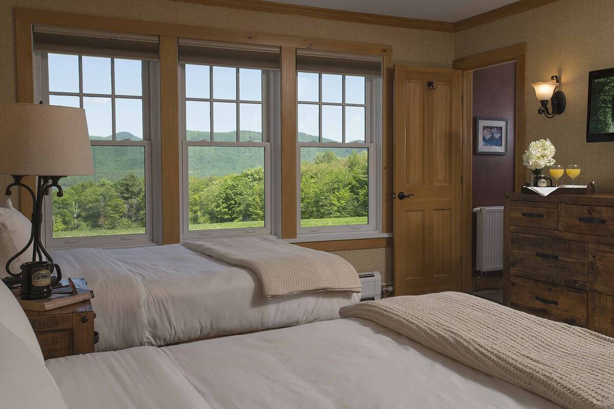Un dormitorio con dos camas, ventanas amplias, un aparador, una lámpara y un cuadro en la pared.