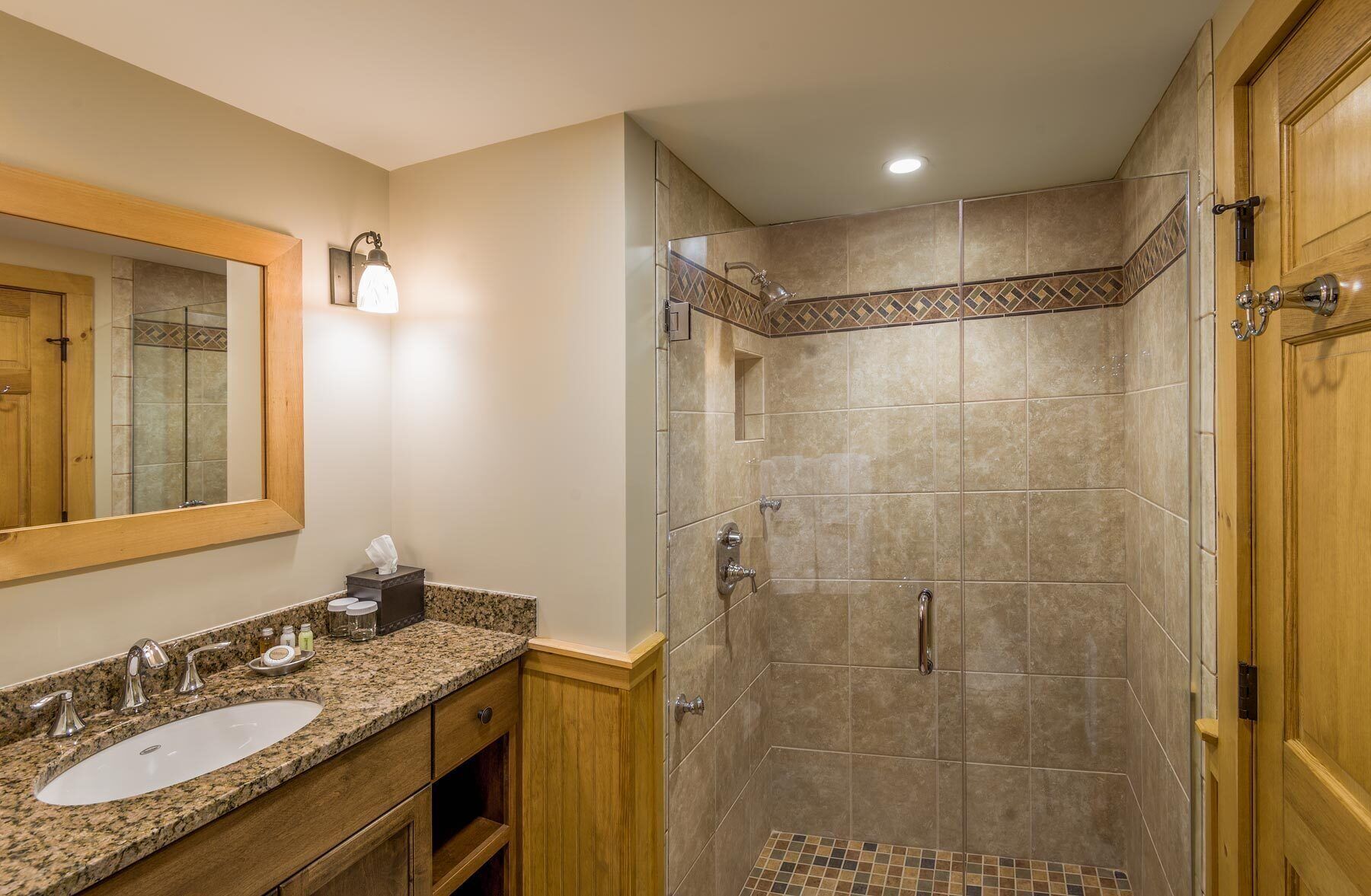 Un baño con lavamanos de encimera de granito, espejo, dispensador de toallas y ducha con suelo de azulejos.