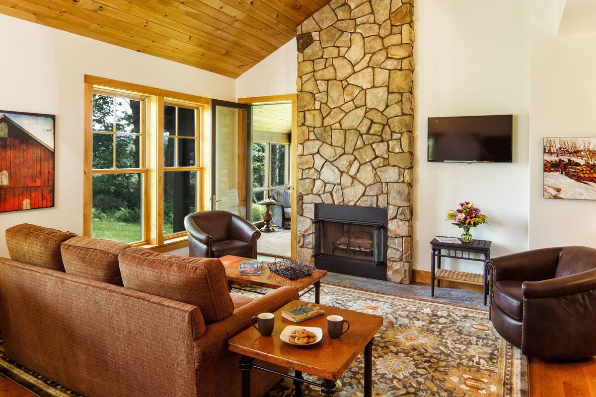 Un salón acogedor con chimenea de piedra, sofás de cuero y un amplio ventanal con vistas a un jardín.