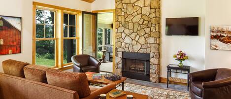 A cozy living room with a stone fireplace, leather sofas, and a large window overlooking a garden.