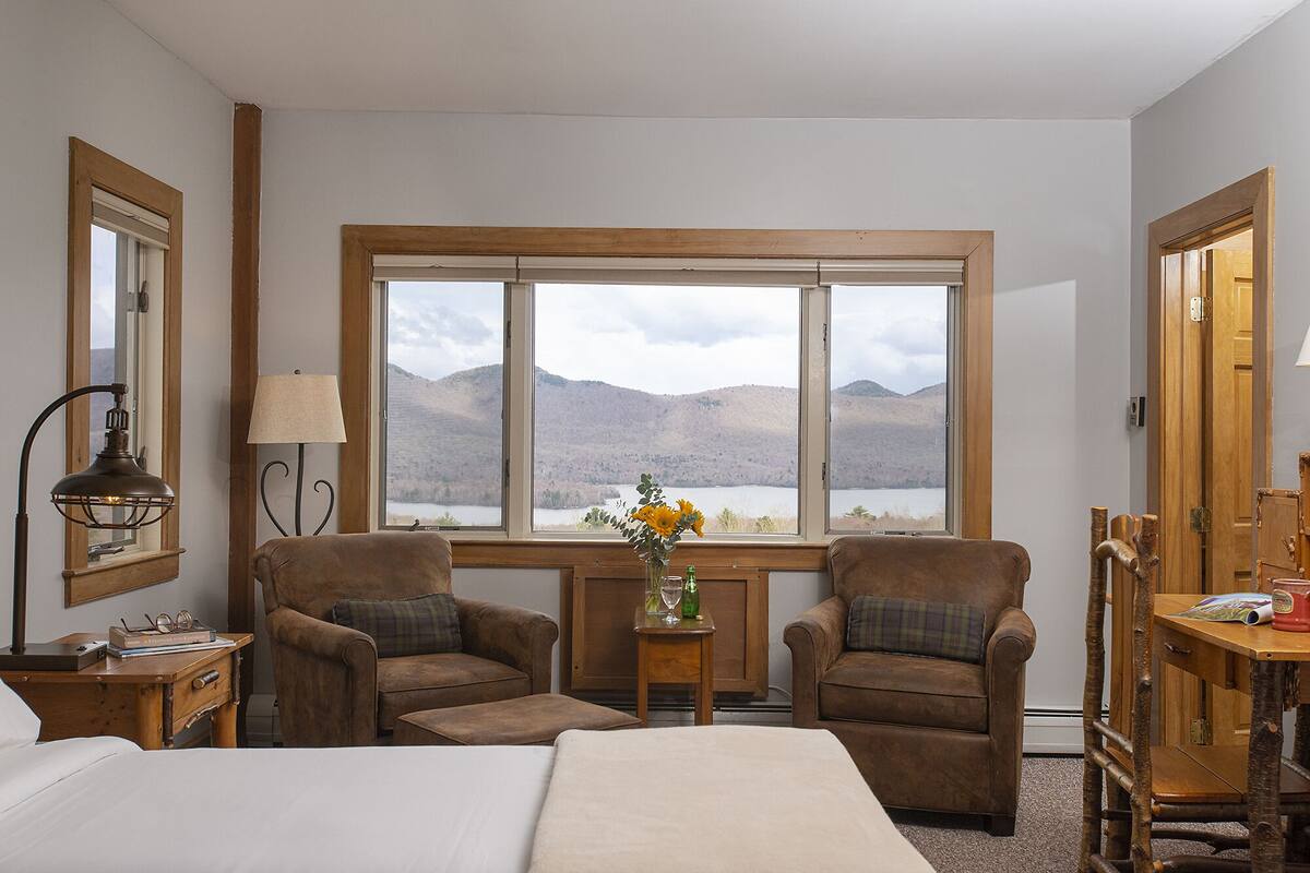 Una habitación con dos sillones de cuero, una cama, una mesita de noche, una lámpara, una pequeña mesa con un jarrón de flores y un ventanal con vistas a la montaña.