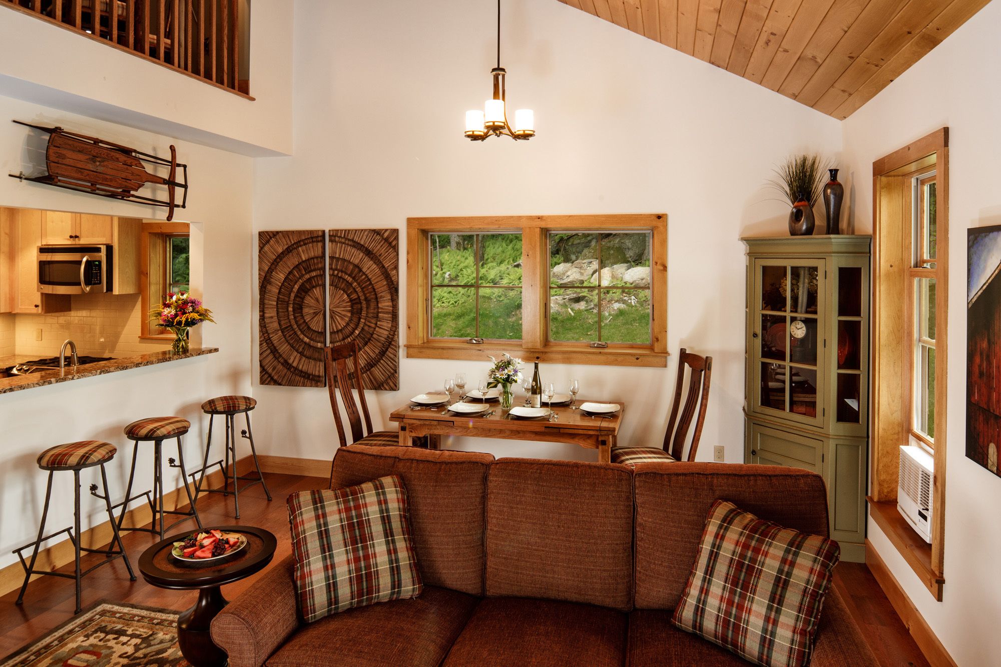 A cozy living room with a brown sofa, plaid cushions, a dining table set for four, and a large window with a view of greenery.