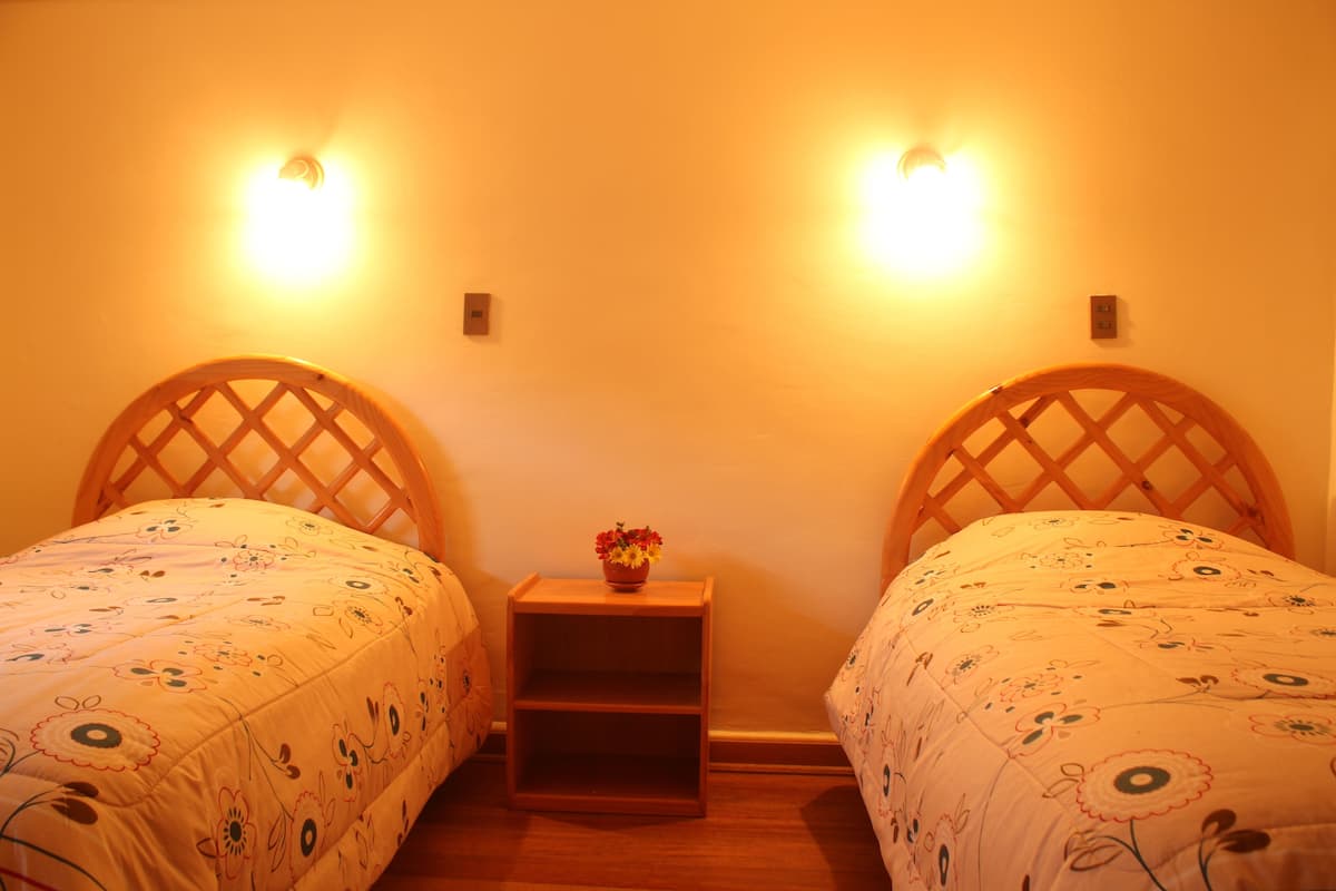 Una habitación con dos camas de madera, cada una con una colcha de estampado floral, una mesita de noche de madera con un jarrón de flores, y lámparas fijadas a la pared.
