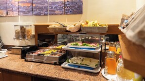 A buffet with various food items and beverages, including fruits, salads, and hot dishes, displayed on a counter with a cityscape view in the background. - Hotel Angelica (Florence)