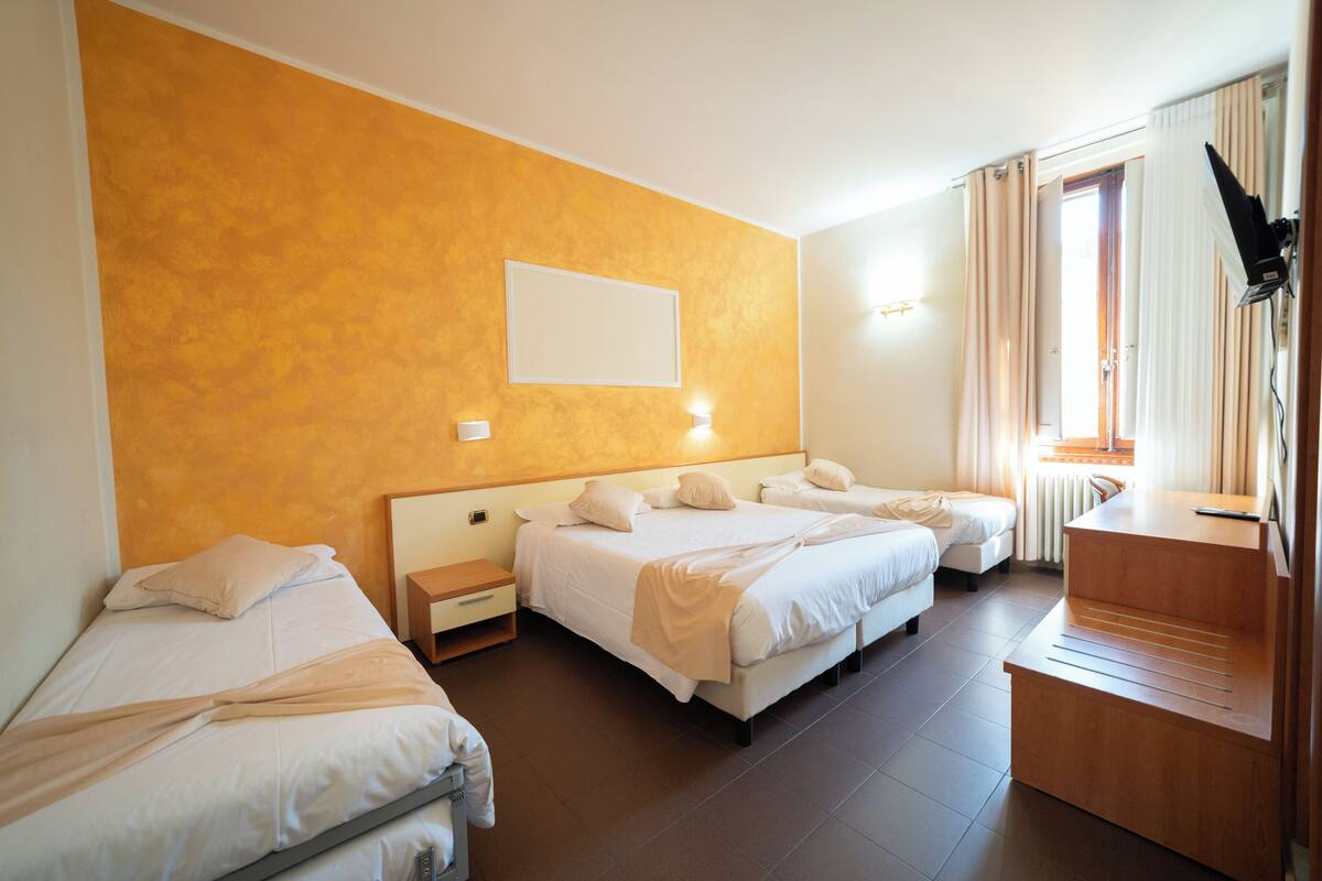 A hotel room with two single beds and a double bed, a small bedside table, a built-in desk, a chair, and a large window with curtains.