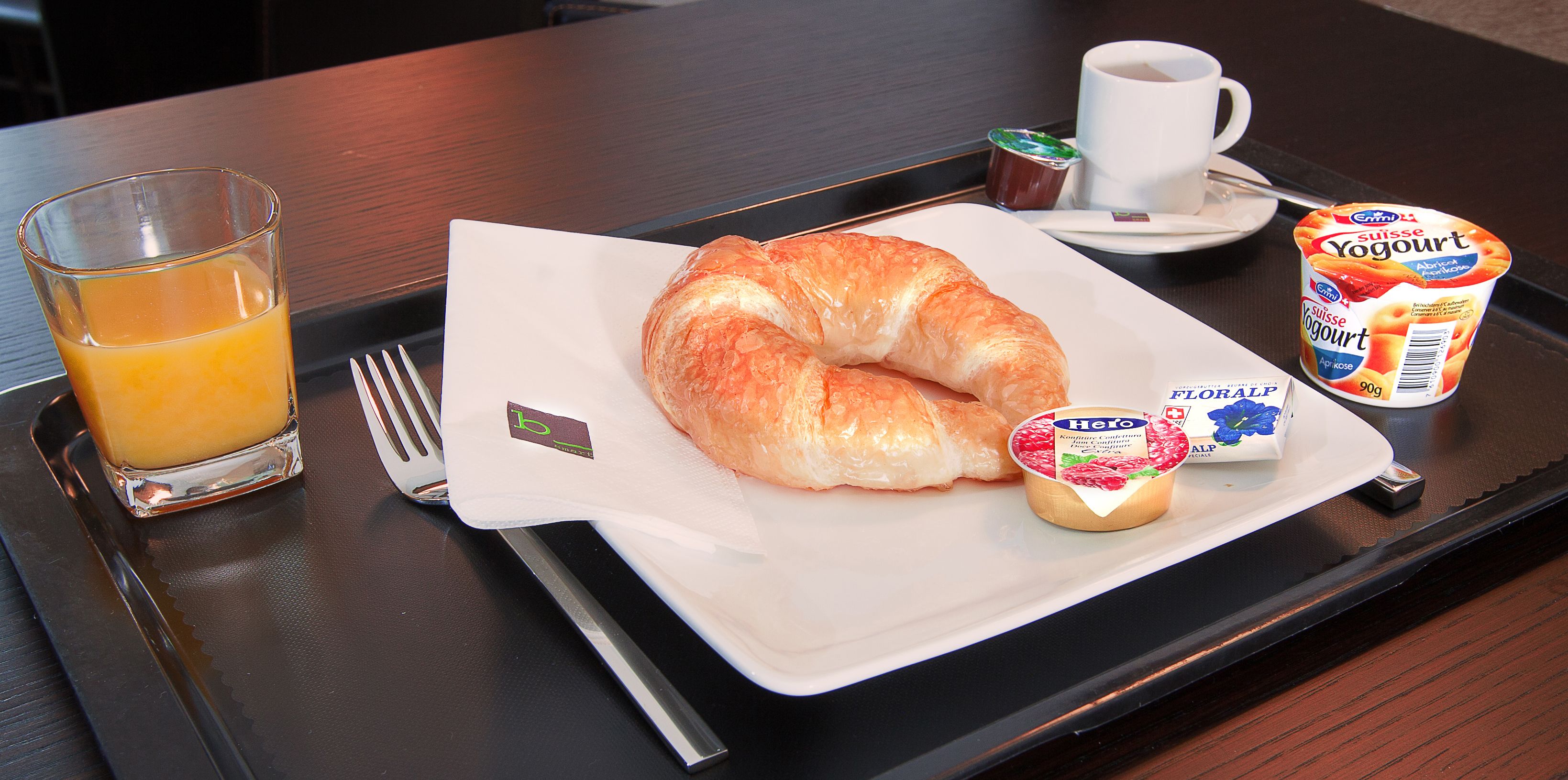 Un plateau de petit-déjeuner avec un croissant, un verre de jus d’orange, une tasse de café et un pot de yaourt.