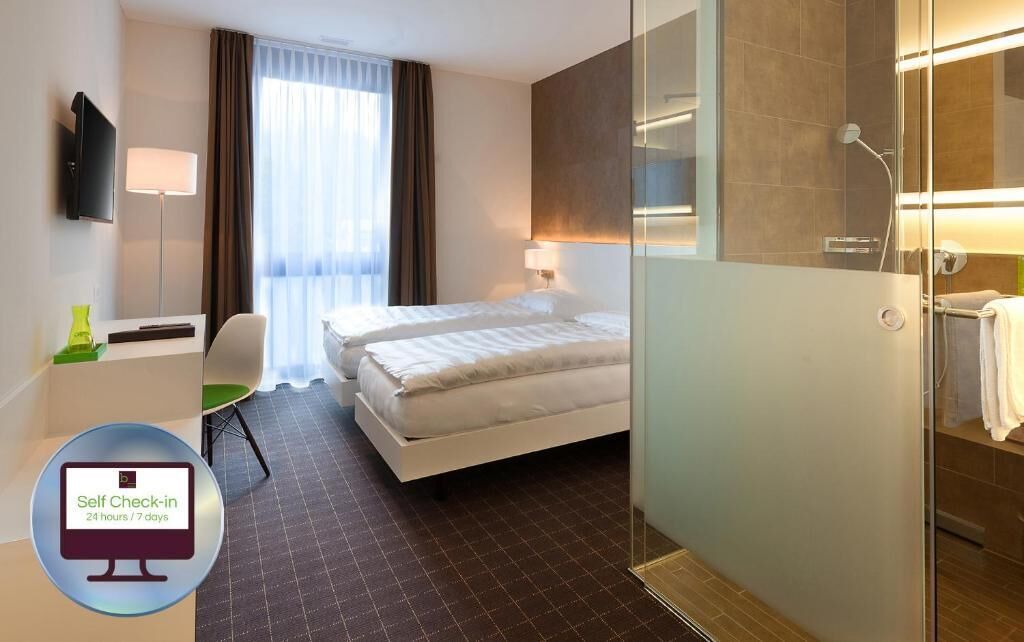 A hotel room with a bed, a desk with a chair, a TV, and a self-check-in kiosk.
