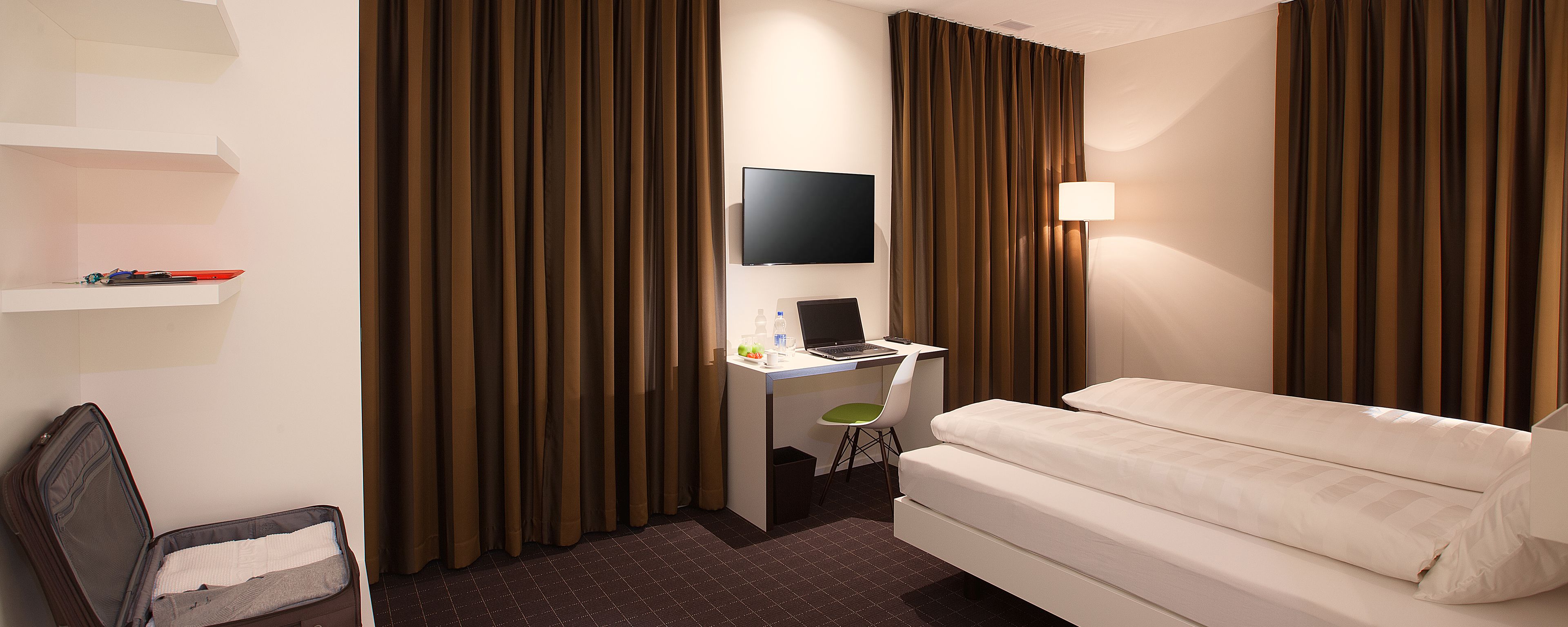 A hotel room with a bed, a desk with a laptop, a TV, and curtains.