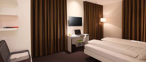 A hotel room with a bed, a desk with a laptop, a TV, and curtains.