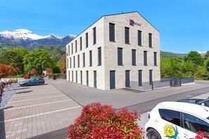 Un edificio moderno con un letrero 'b hotel', un estacionamiento y montañas al fondo.